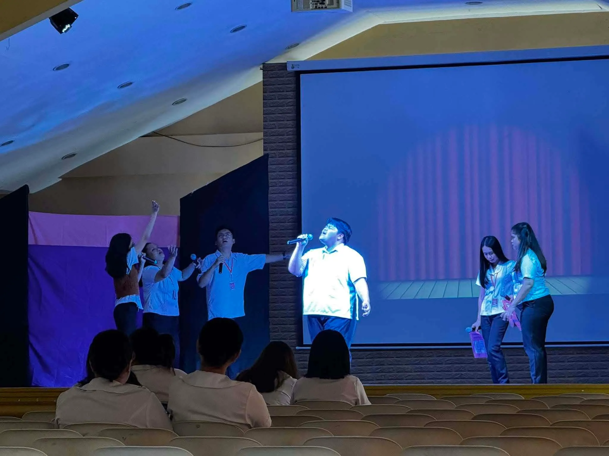 Performers on stage under blue light, one singing in front of the screen.
