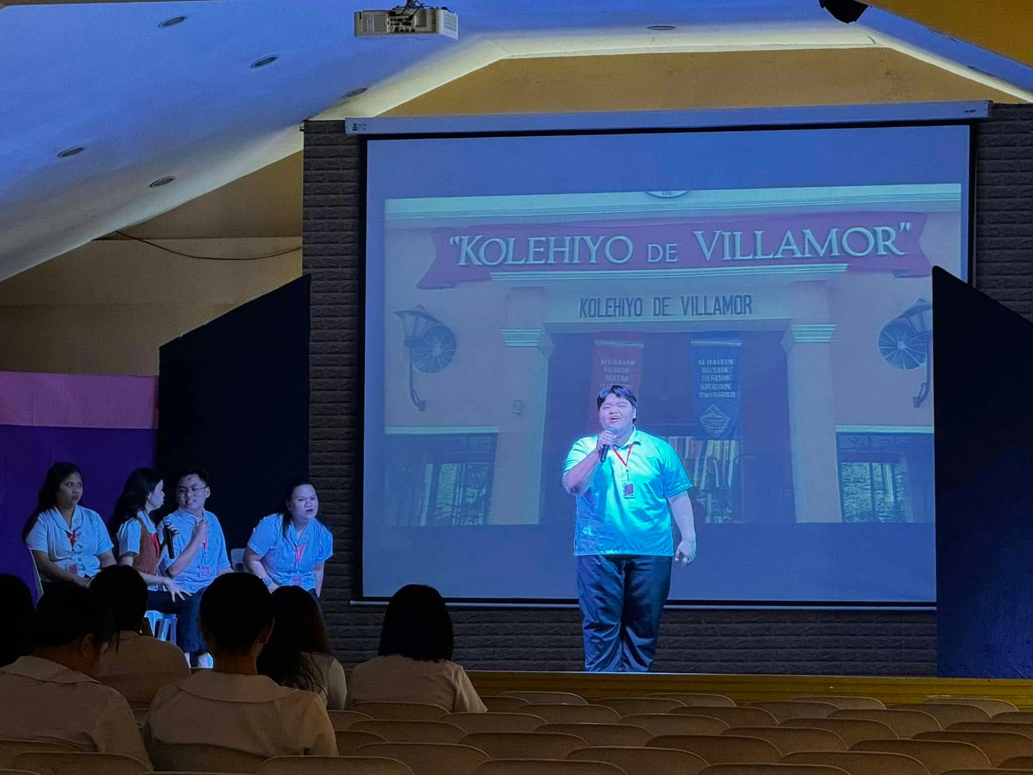 A performer at center stage in front of the “Kolehiyo de Villamor” title screen.