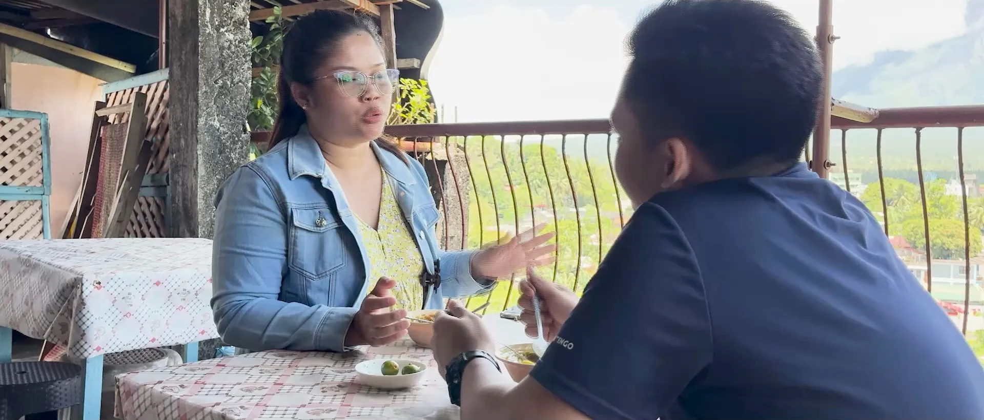 A still from Balik-Tanaw, a woman in a denim jacket talking with a young man over a meal on a veranda, mountains behind them.